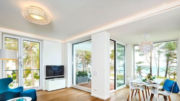 A modern living room with a flat-screen TV, a dining area with chairs, and large windows offering a view of the outdoors. - Villa Vogue, Austernfischer (Binz)