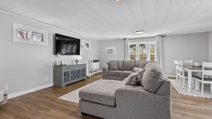 Olohuoneessa on harmaa sohva, litteänäyttöinen televisio, harmaa kaappi ja ruokailutila, jossa on valkoiset huonekalut.