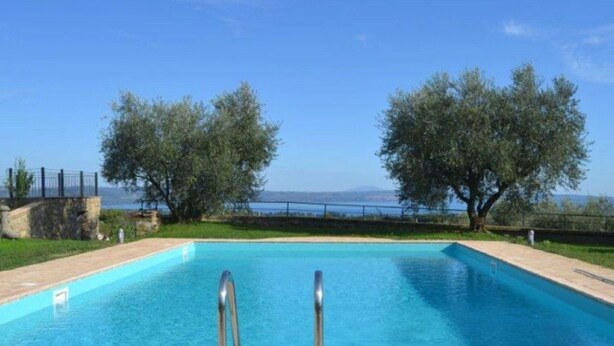 A rectangular swimming pool with metal ladders on both sides, surrounded by a stone wall and a grassy area with trees. - Belvilla by OYO Villa Degli Olivi (Grotte di Castro)