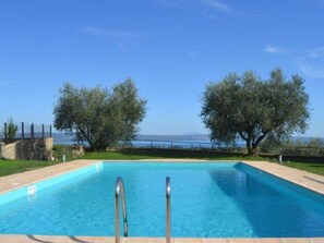 A rectangular swimming pool with metal ladders on both sides, surrounded by a stone wall and a grassy area with trees. - Belvilla by OYO Villa Degli Olivi (Grotte di Castro)