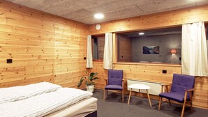 Una habitación con paredes de madera, una cama, dos sillas moradas, una mesita y una ventana con cortinas.