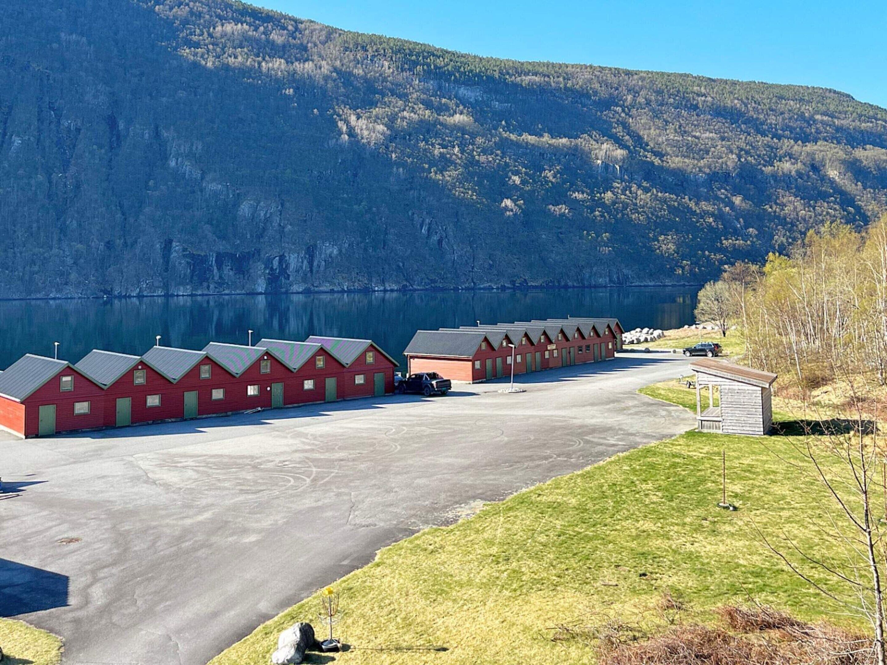 A row of red cabins with a parking area in front, set against a backdrop of a lake and mountains.