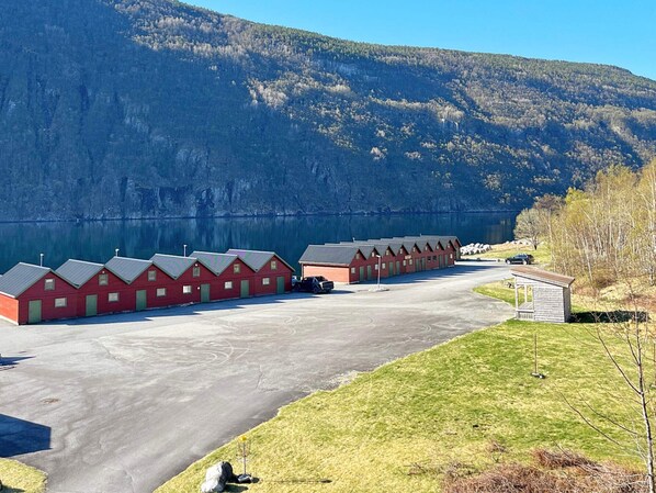 A row of red cabins with a parking area in front, set against a backdrop of a lake and mountains.