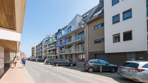 A multi-story residential building with balconies, parked cars, and a clear sky.