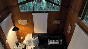 A cozy cabin with a black leather sofa, a ceiling fan, and a window with a view of bamboo. - HW Tiny Home Cidra (Cidra)
