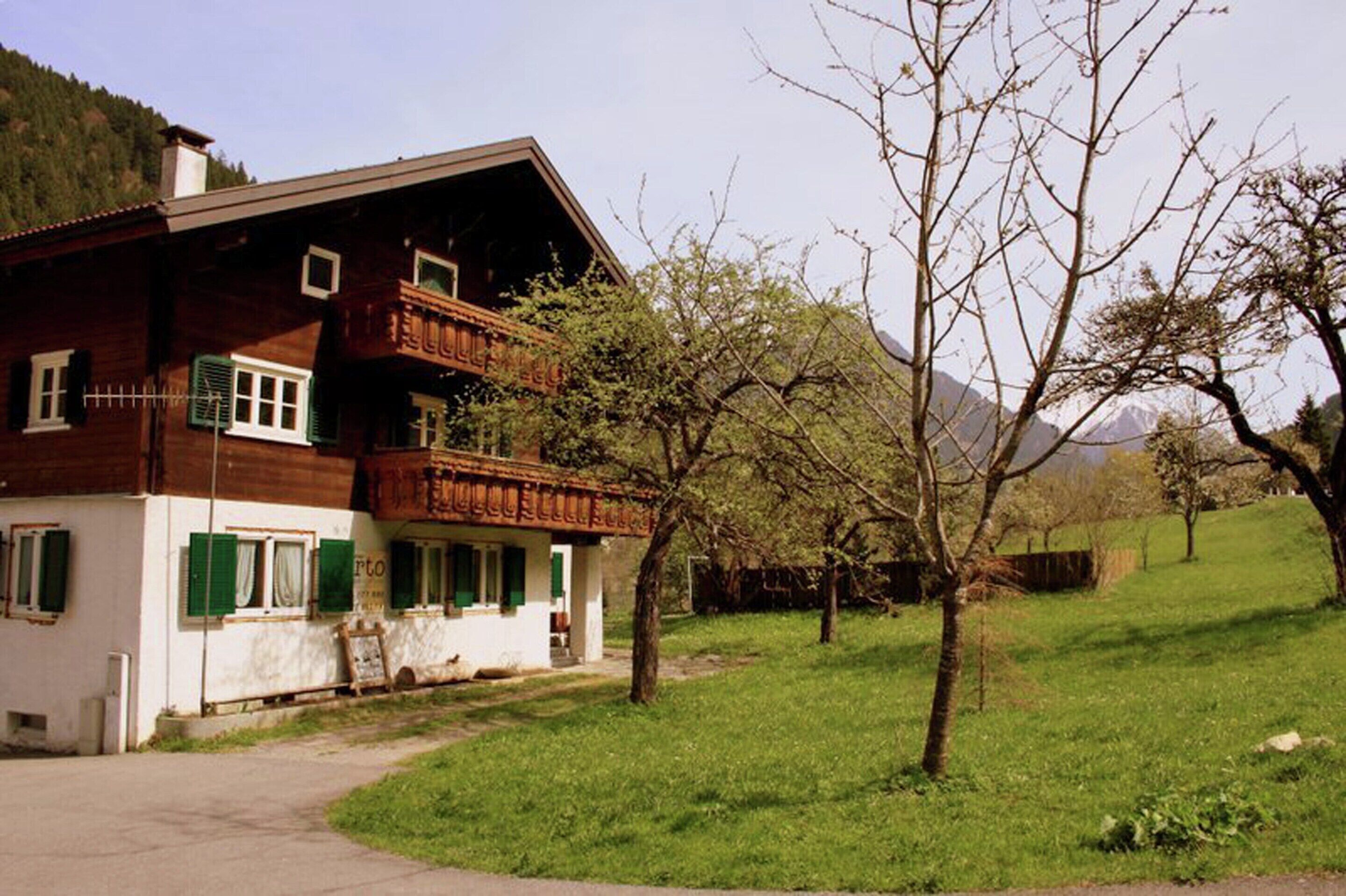 Une maison en bois avec un balcon, une cour verdoyante et une montagne en arrière-plan.