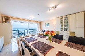 Un espace de repas moderne avec une grande table, des chaises et un vase de fleurs. Une grande fenêtre offre une vue sur l’océan.