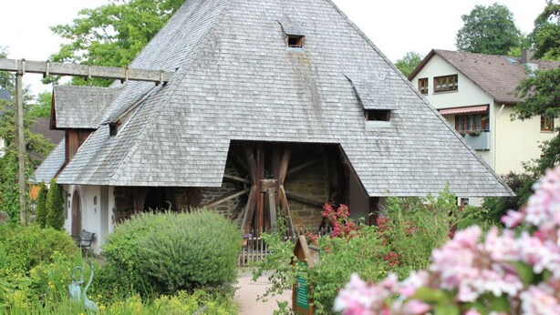 A traditional wooden house with a large water wheel attached to the roof, surrounded by lush greenery and a garden path. - Holiday Apartment Holzwieb (Unterkirnach)