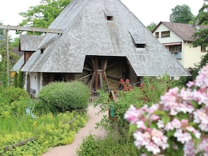 Una casa de madera tradicional con una noria grande unida al tejado, rodeada de vegetación exuberante y un sendero en el jardín.