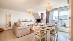 A modern living room with a sofa, dining table, and chairs, a kitchen area with a TV, and large windows offering a view of the outside. - Coastal Retreat With Balcony (Koksijde)