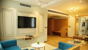 A modern hotel room with a flat-screen TV, blue sofas, a small round table with a plant, and a wooden reception desk. - Cosmos Westend City Centre (Udaipur)