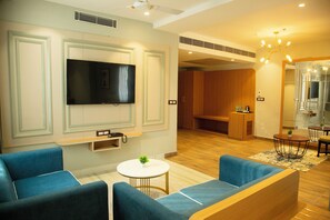 A modern hotel room with a flat-screen TV, blue sofas, a small round table with a plant, and a wooden reception desk. - Cosmos Westend City Centre (Udaipur)