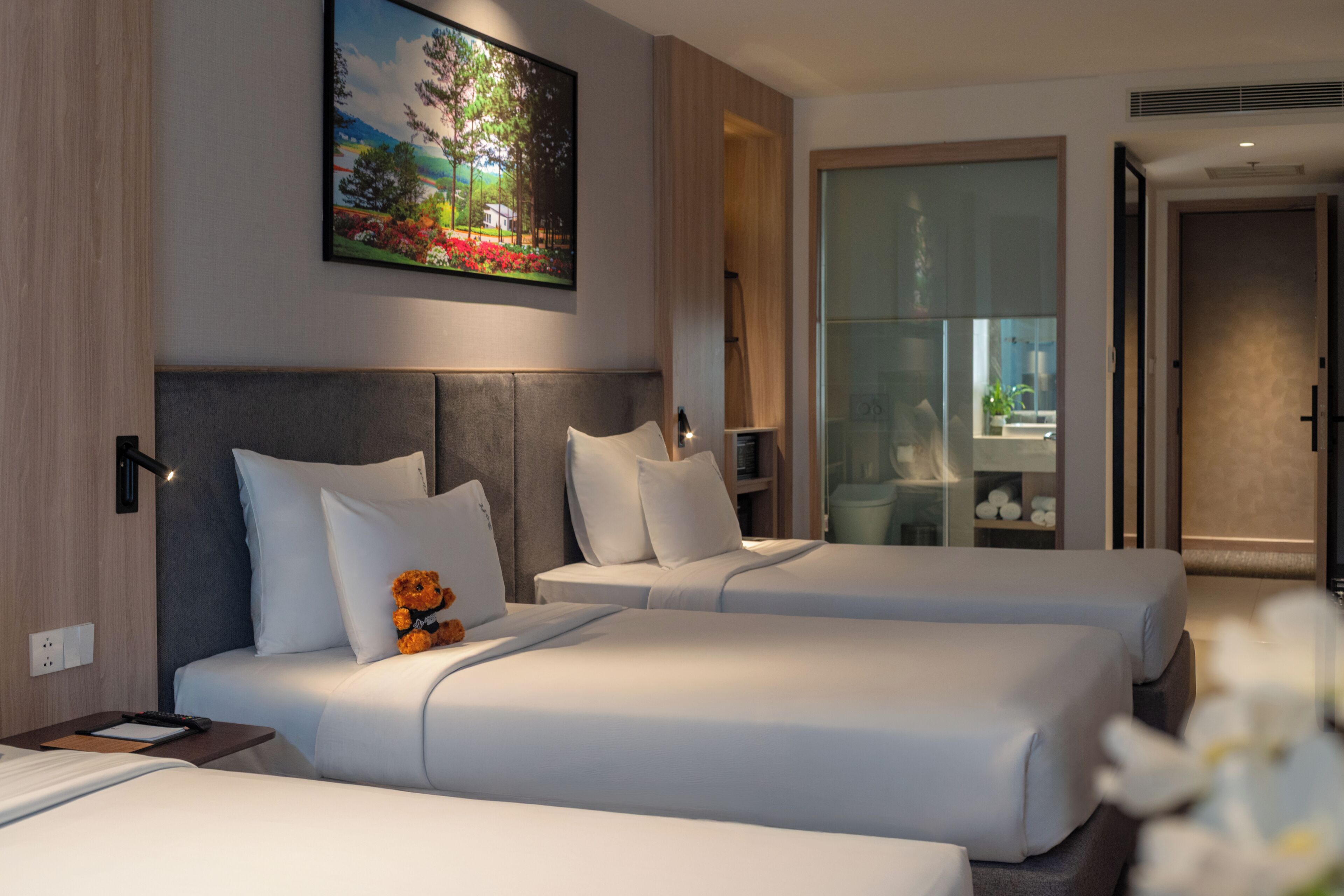 A hotel room with two beds, a painting on the wall, a nightstand, and a view through a glass door.