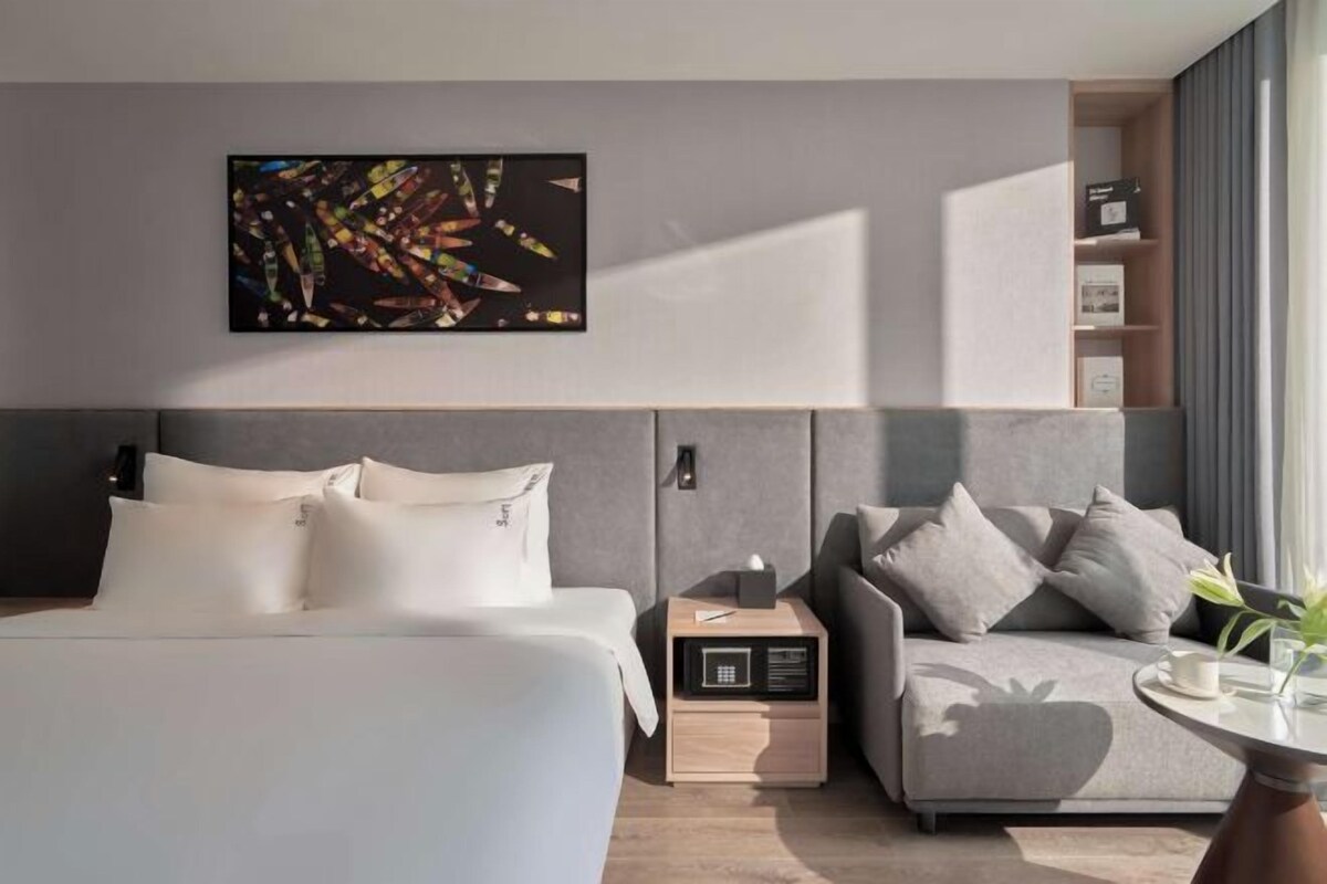 A modern hotel room with a bed, a sofa, a bedside table, a wall-mounted artwork, and a built-in shelf with books.