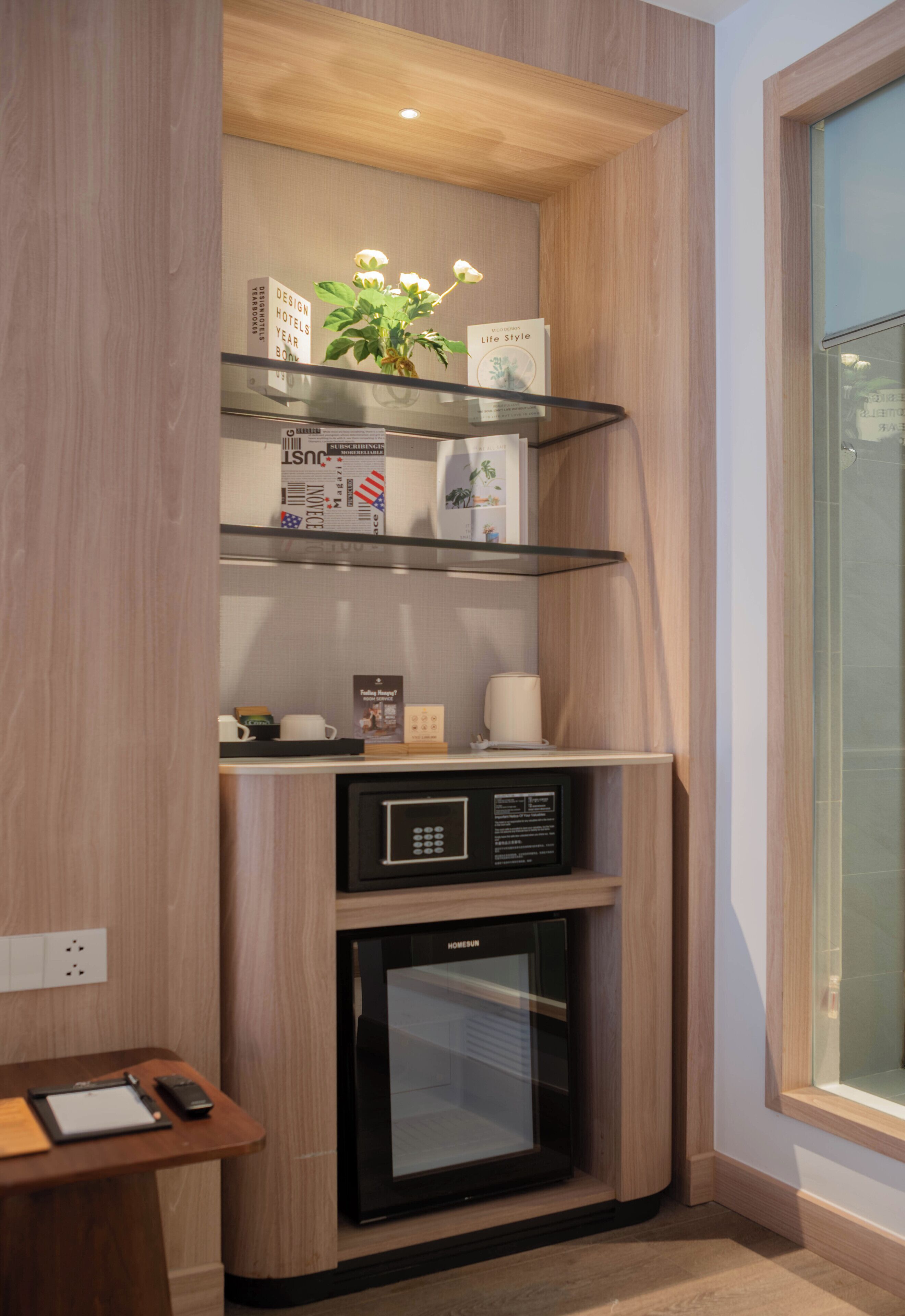 A corner kitchenette with a built-in microwave, a small refrigerator, and a shelf with books and a plant.
