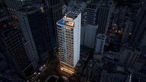 A high-rise building with a prominent 'ADAMAS' sign on the top. It features multiple floors with balconies, and the lower part of the building is illuminated, suggesting interior lighting.