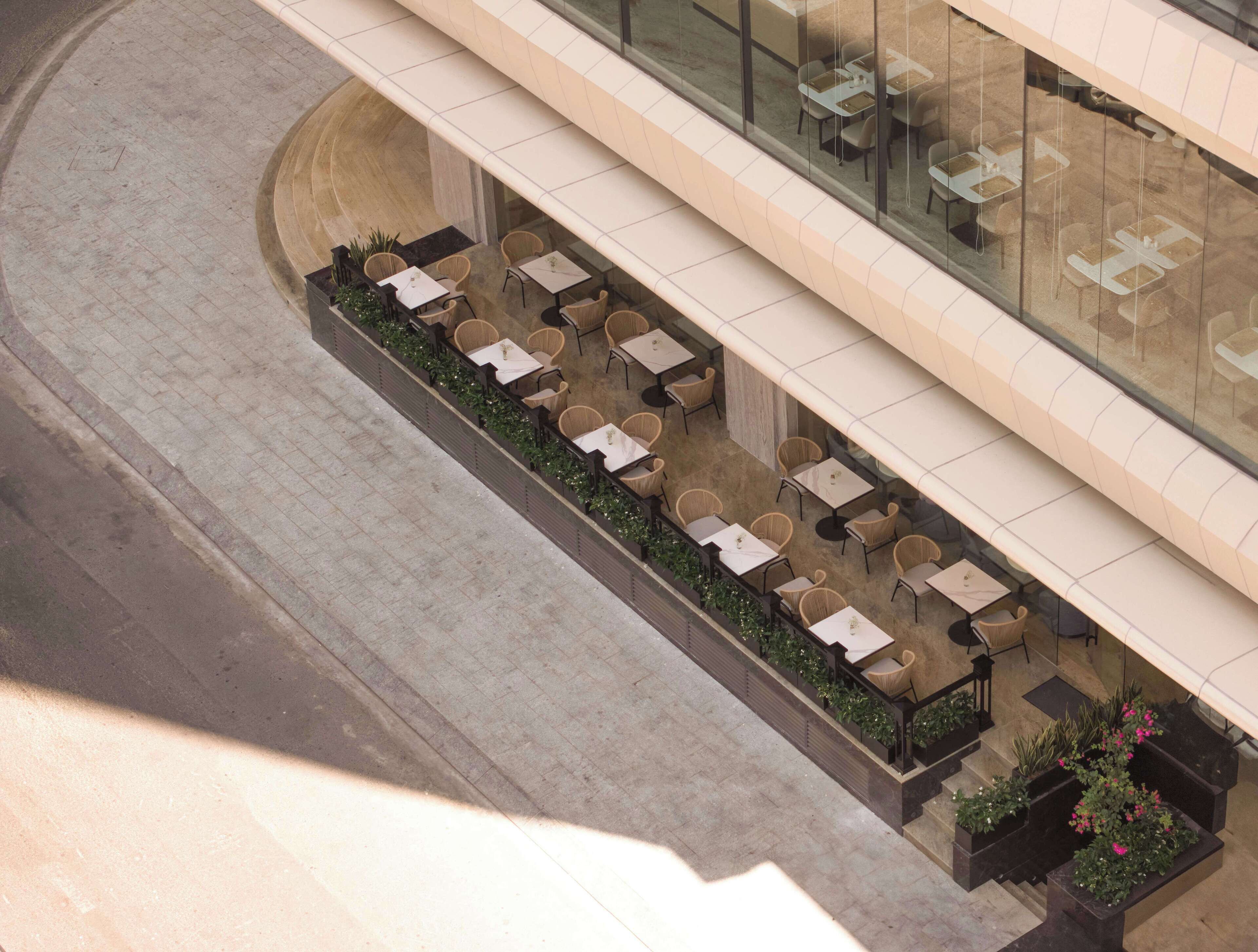 An outdoor seating area with tables and chairs, surrounded by greenery, and adjacent to a building with a glass facade.