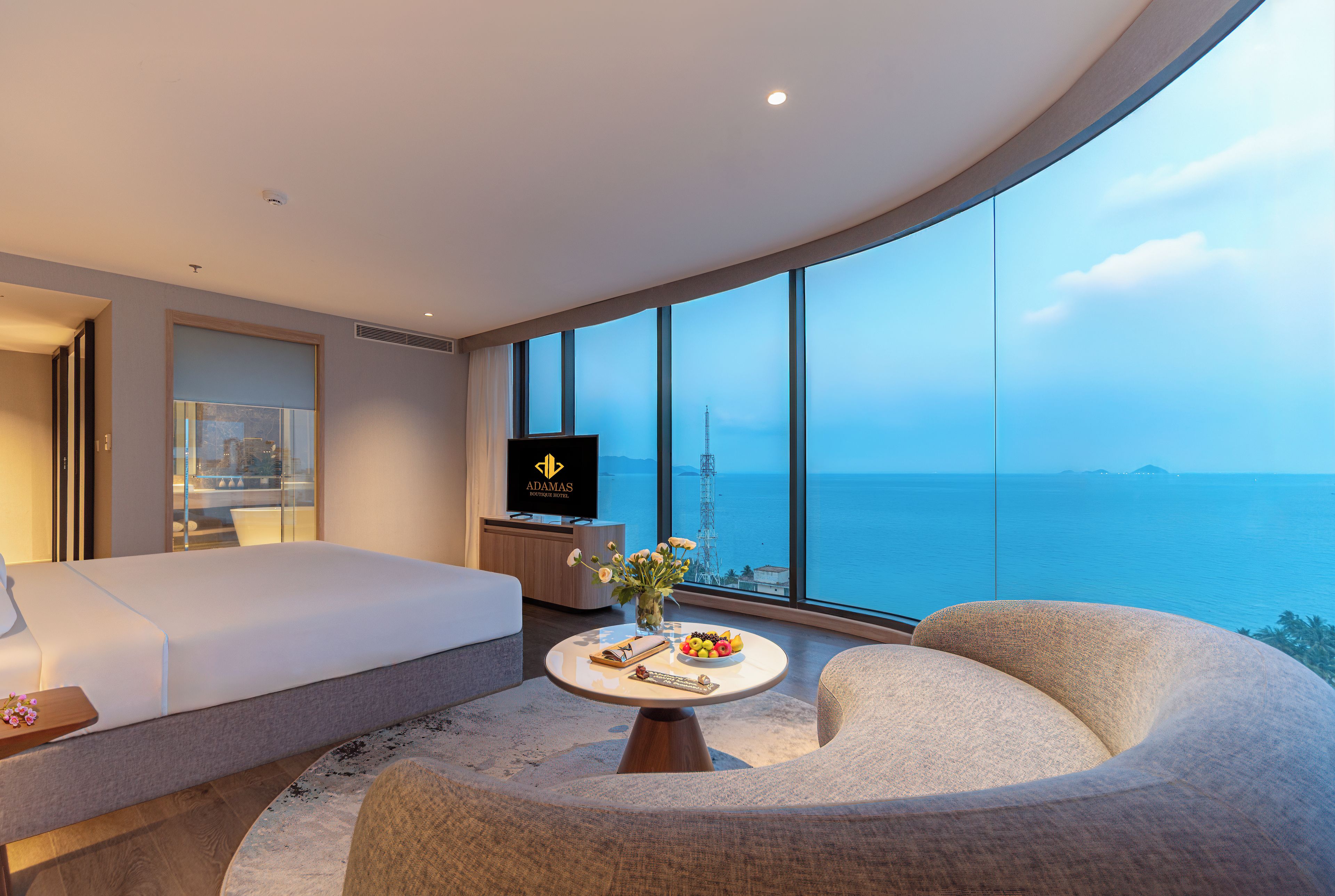 A modern hotel room with a large bed, a flat-screen TV, a dining table with fruit and a vase, and a view of the sea.