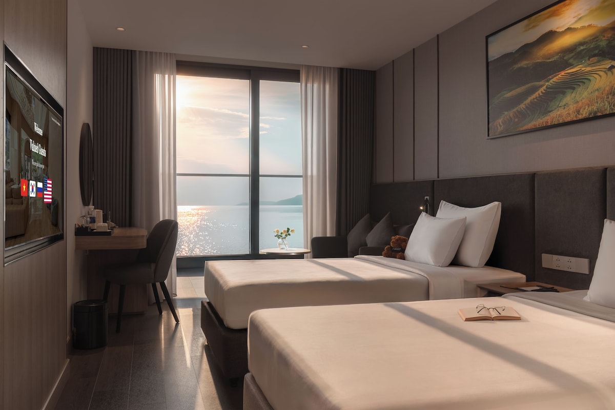 A hotel room with two beds, a desk, a chair, a TV, and a large window with a view of the water.