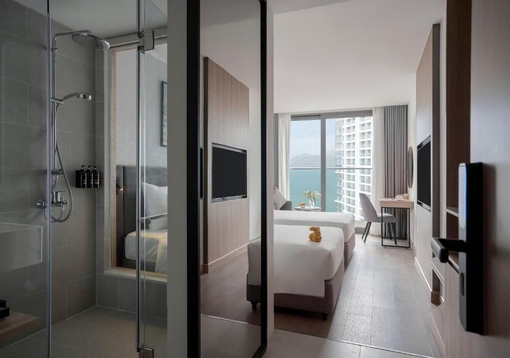A hotel room with a glass-enclosed shower, a bed, a desk, and a view of a building.