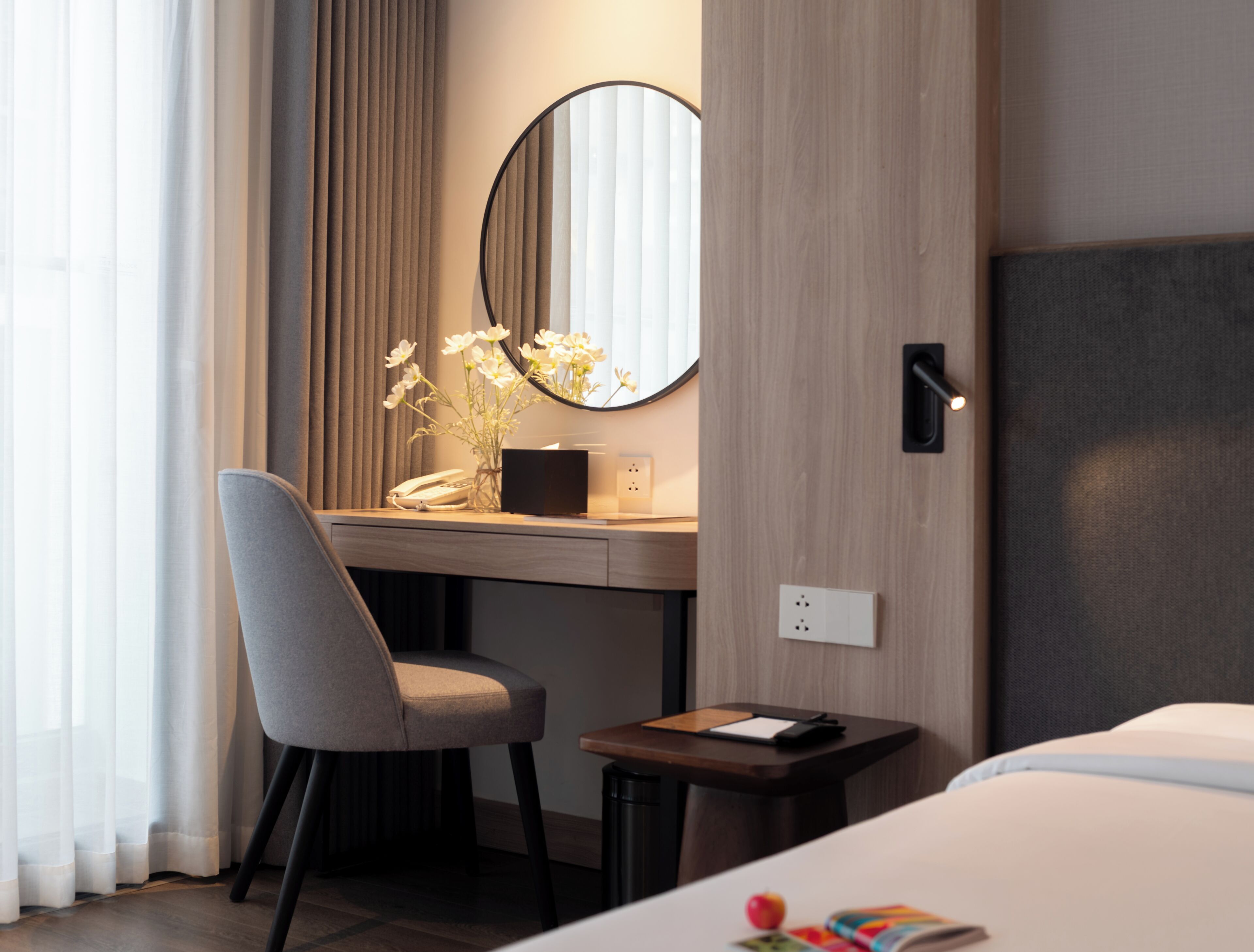 A hotel room with a bed, a desk with a chair, a small table, a mirror, and a vase with flowers.