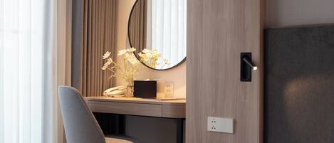 A hotel room with a bed, a desk with a chair, a small table, a mirror, and a vase with flowers.