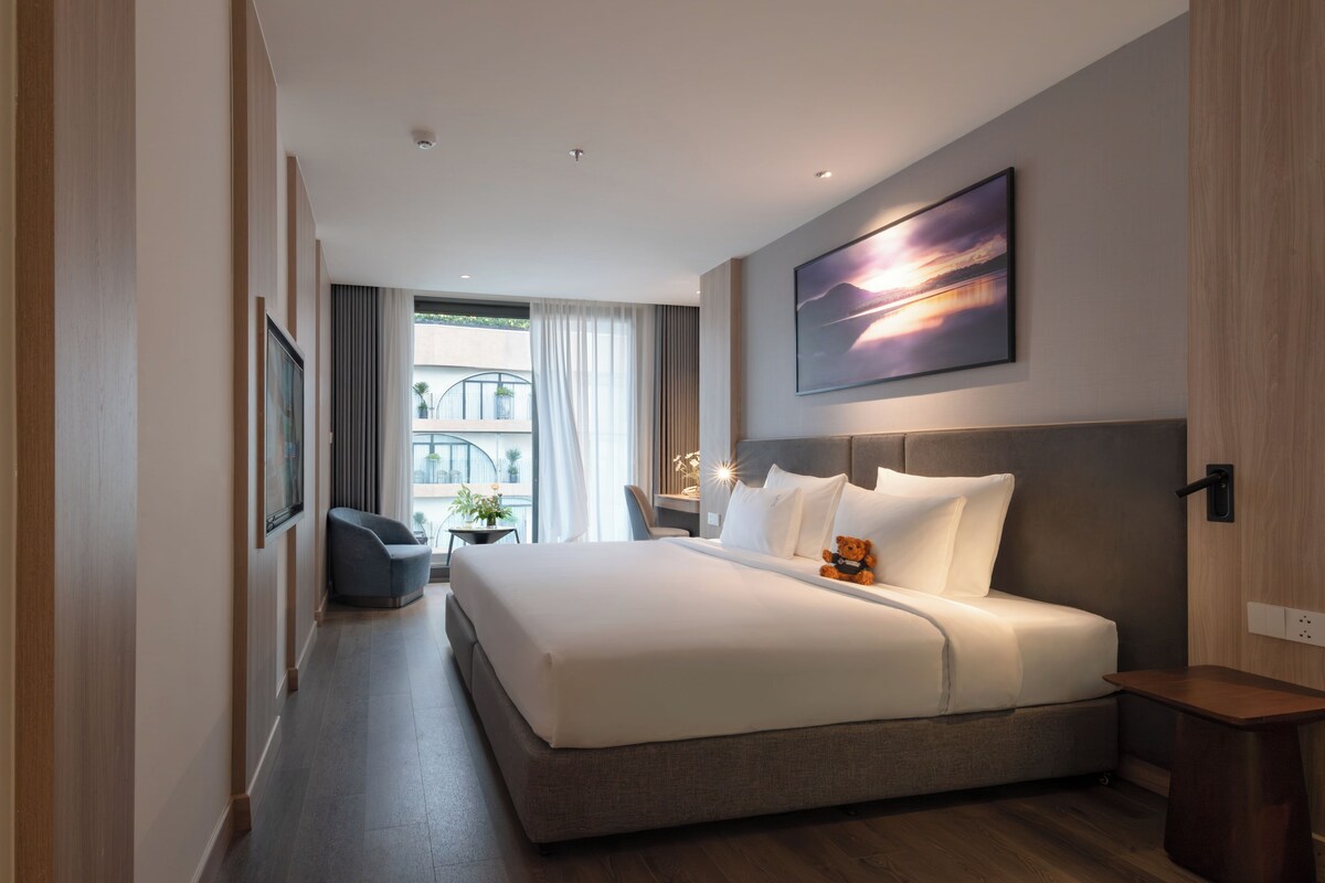 A modern hotel room with a large bed, a desk, a chair, and a view of the building's exterior.