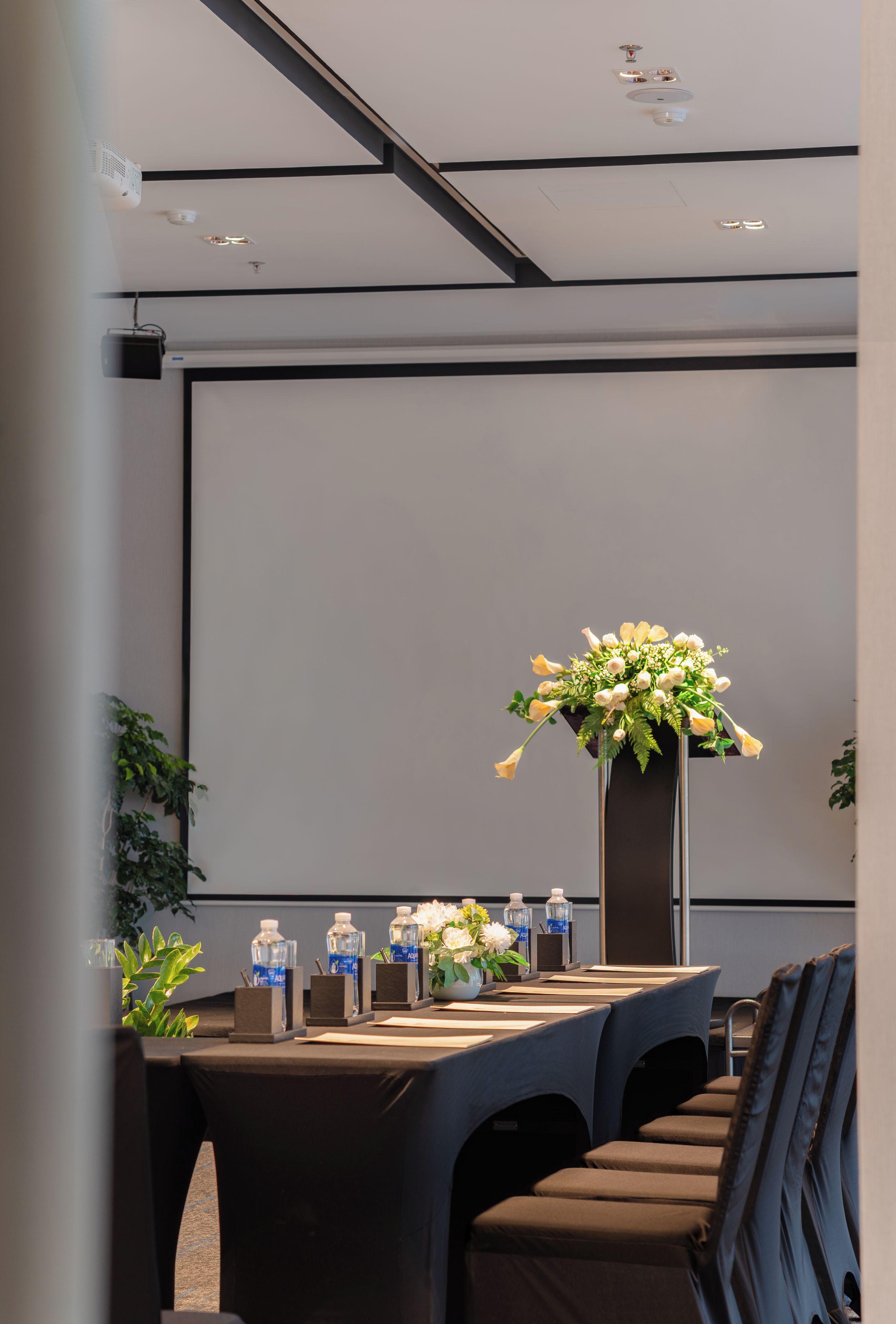 A conference room with a projector screen, a table with water bottles, a vase of flowers, and a seating arrangement.