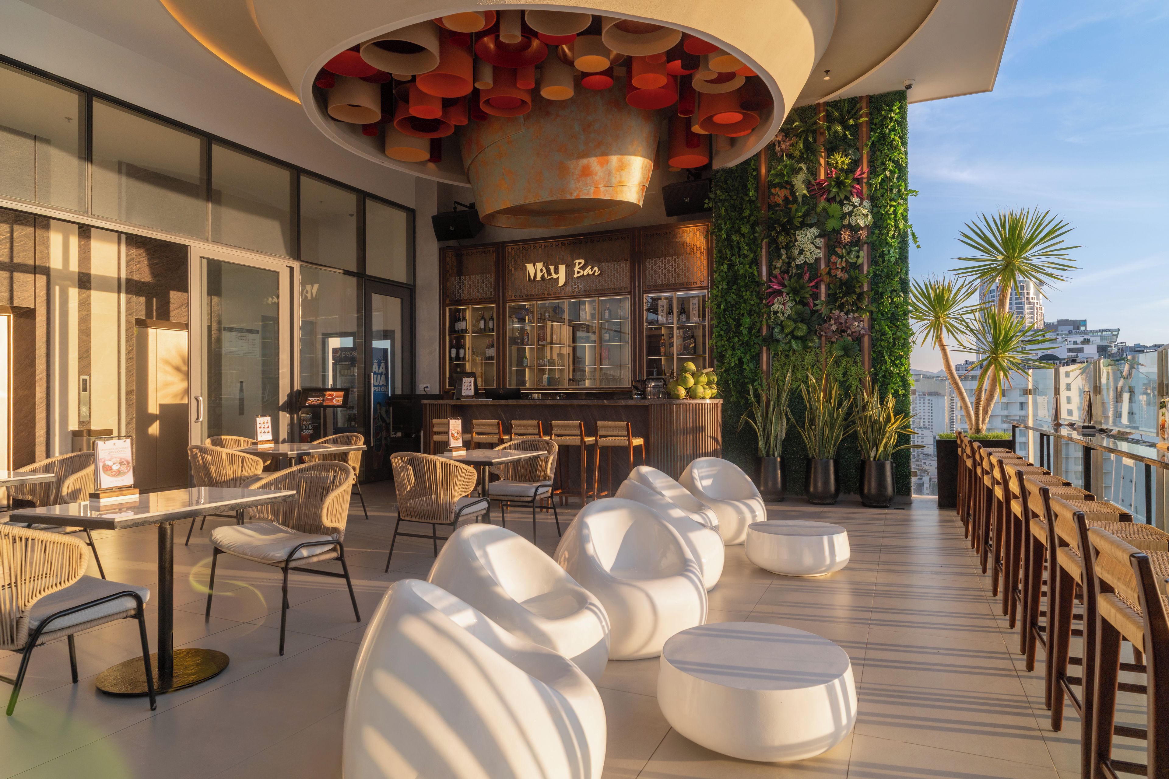 A rooftop bar with outdoor seating, a bar area with a 'May Bar' sign, and a view of the cityscape.