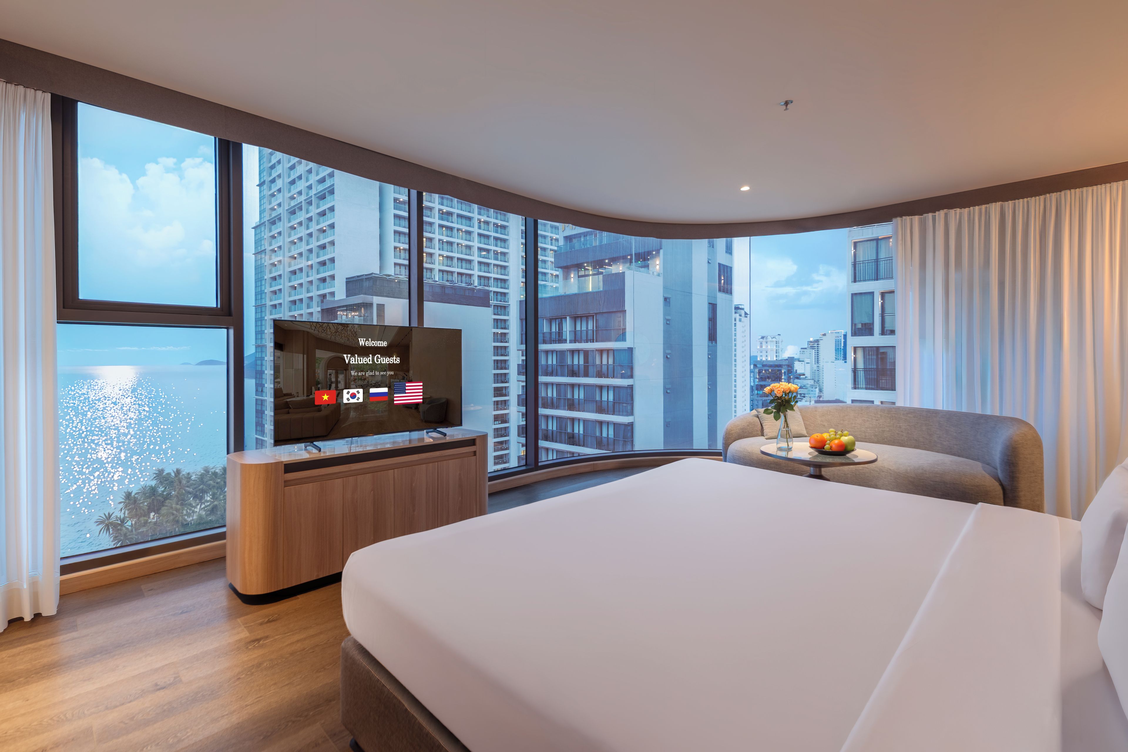 A modern hotel room with a large bed, a TV, a small table with a fruit bowl, and a view of the city and water through large windows.