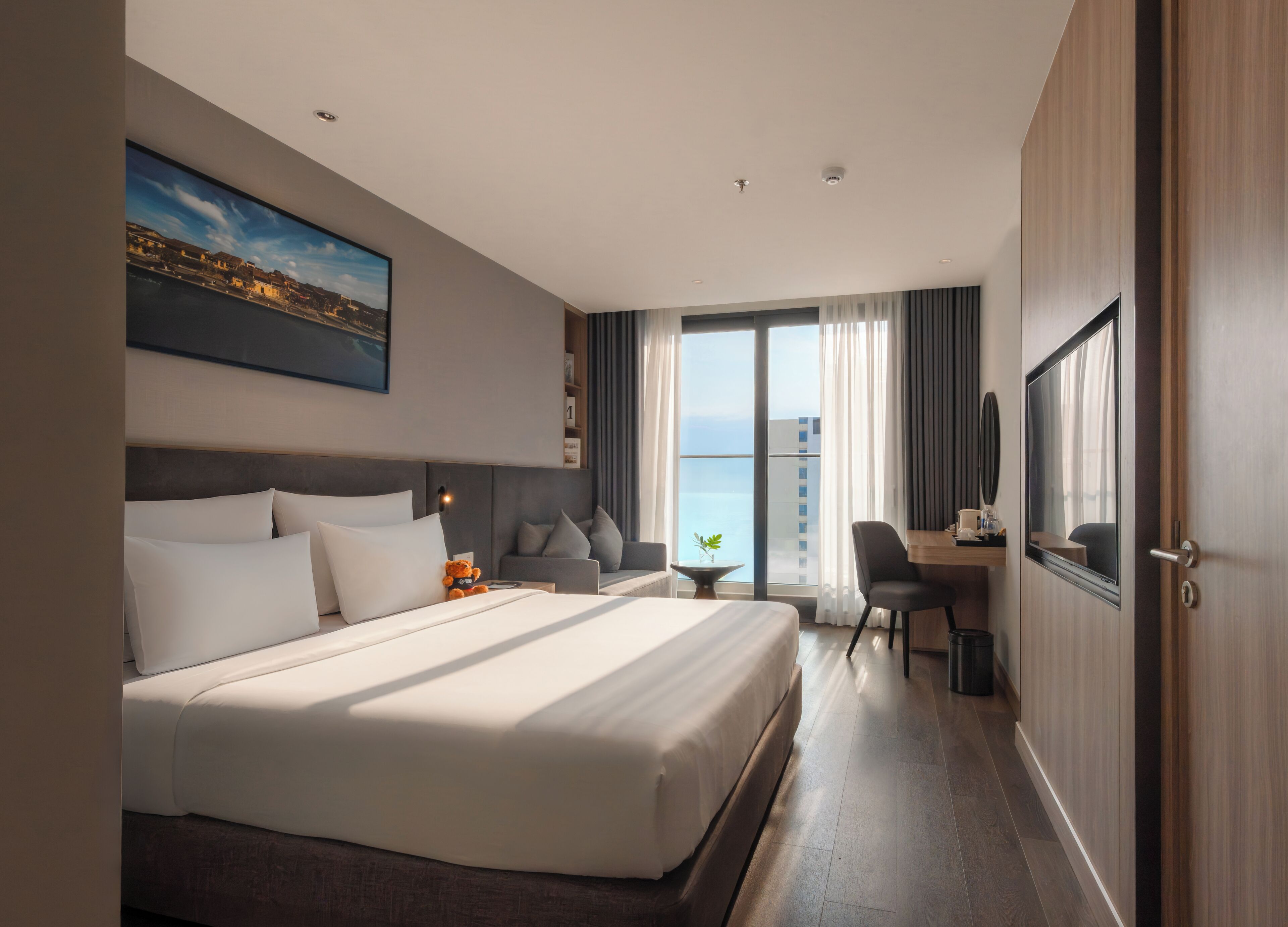 A hotel room with a large bed, a desk, a chair, a TV, and a view of the sea.