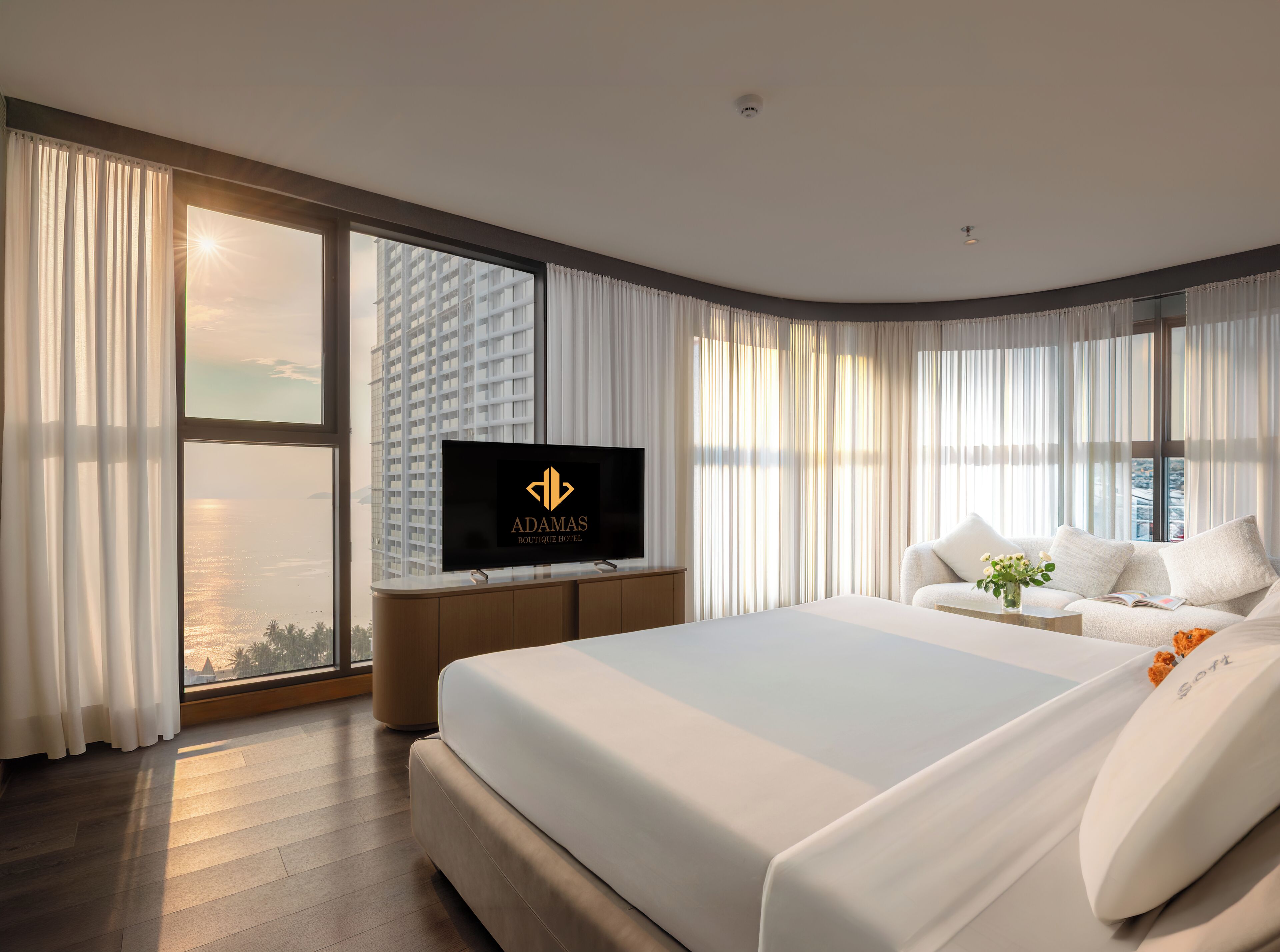 A modern hotel room with a large bed, a flat-screen TV, and a view of a cityscape through large windows.