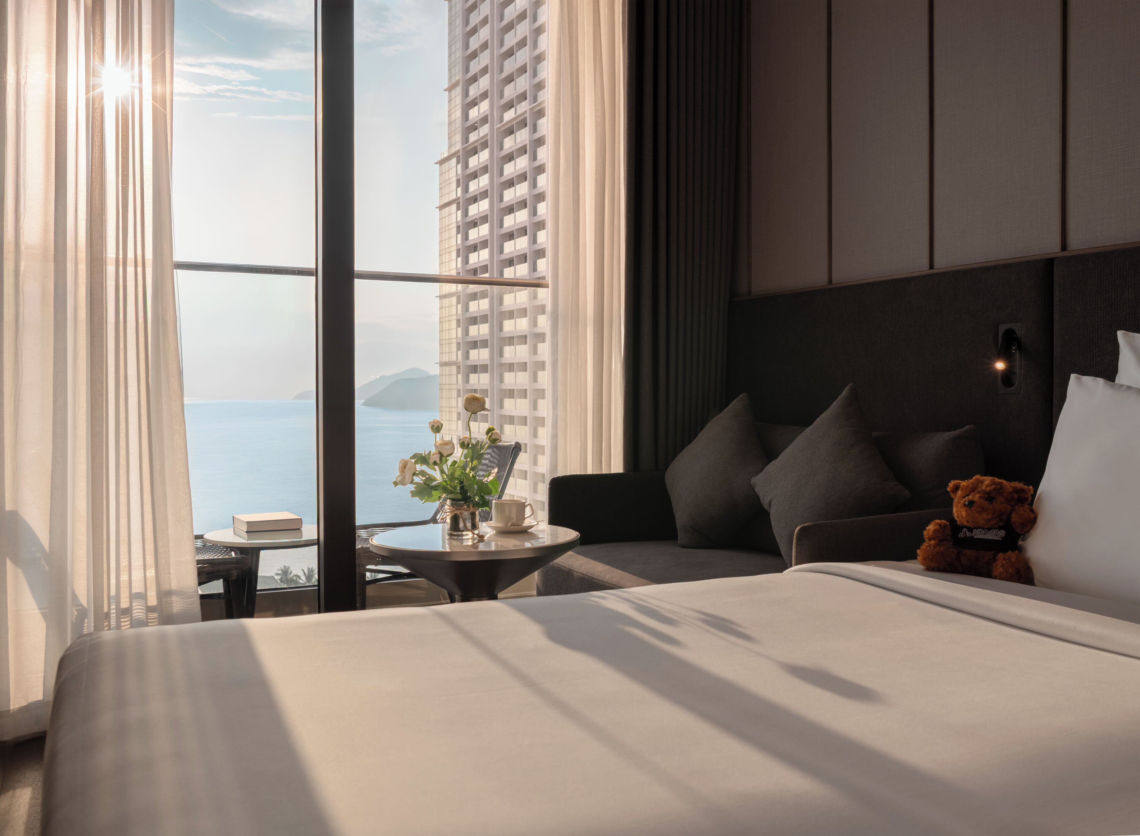 Hotelkamer met een bed, een klein tafeltje met een bloemenvaas, een teddybeer en uitzicht op zee door grote ramen.