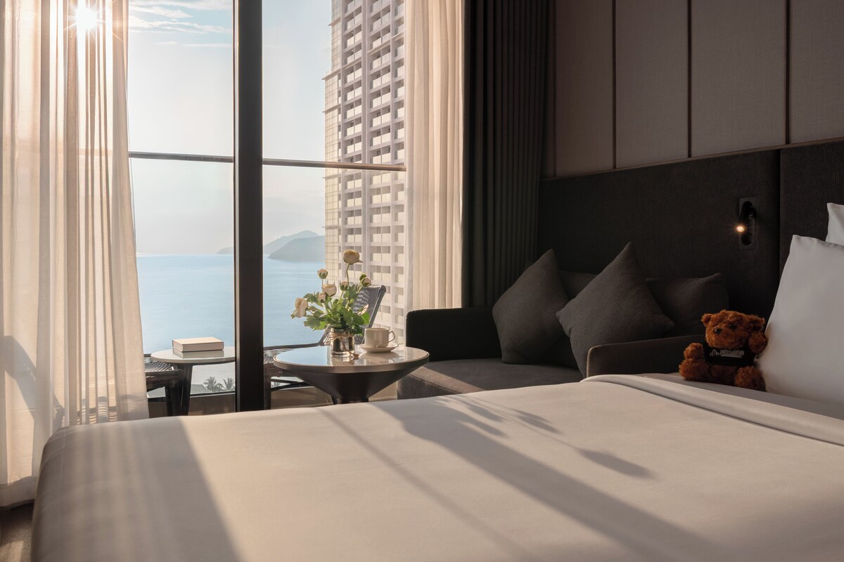 Habitación de hotel con una cama, una mesita con un florero, un osito de peluche y vista al mar a través de ventanas amplias.