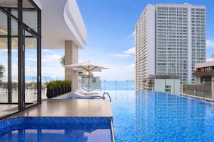 Uma piscina infinita no terraço, vista para o oceano, espreguiçadeiras e um edifício moderno ao fundo.