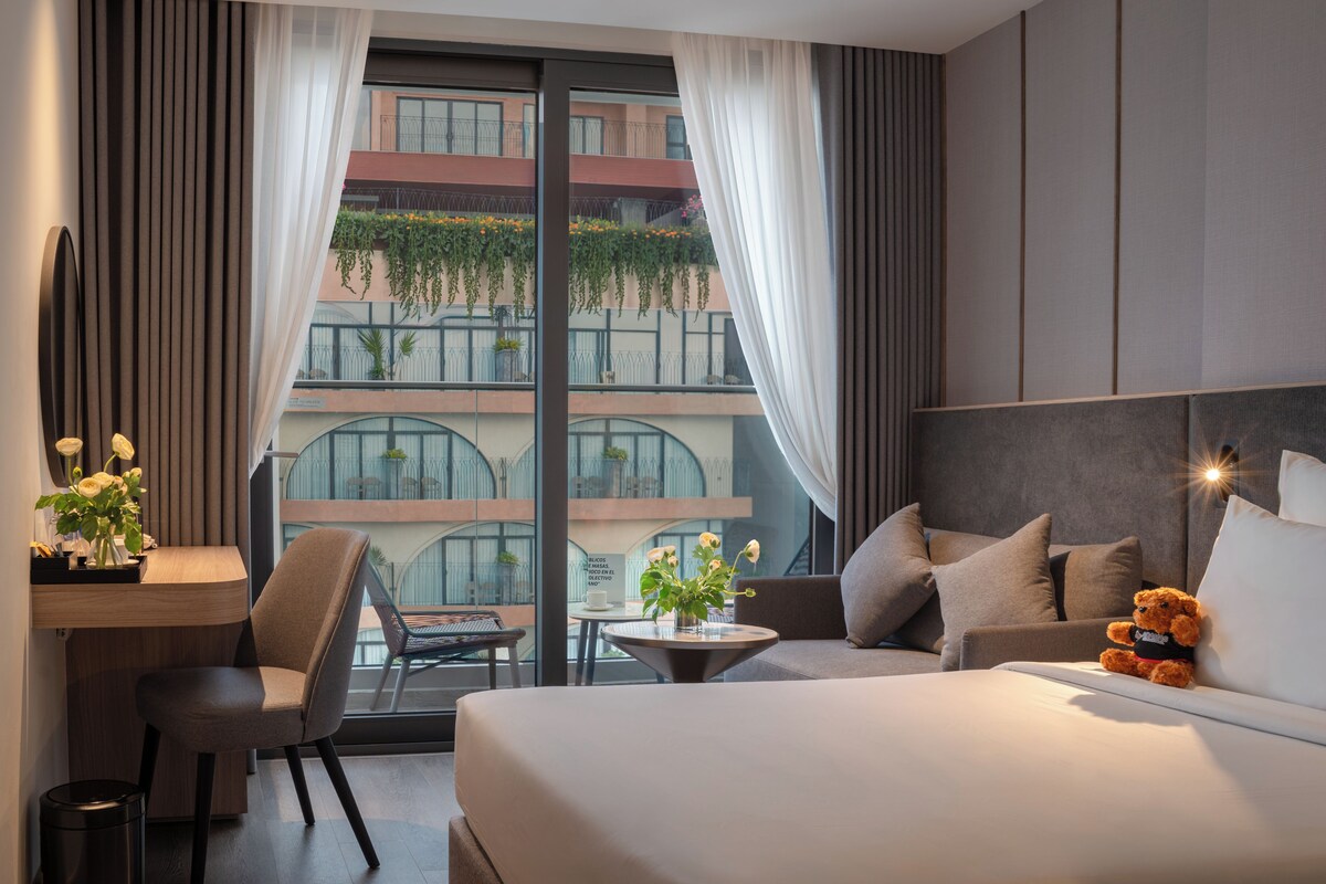 A hotel room with a bed, a desk, a chair, a small table with a vase of flowers, and a view of a building with balconies.