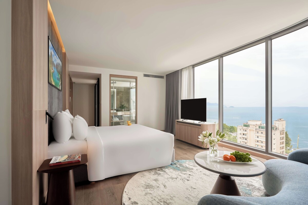 A hotel room with a bed, a TV, a small table with a vase and fruit, and a large window with a city view.