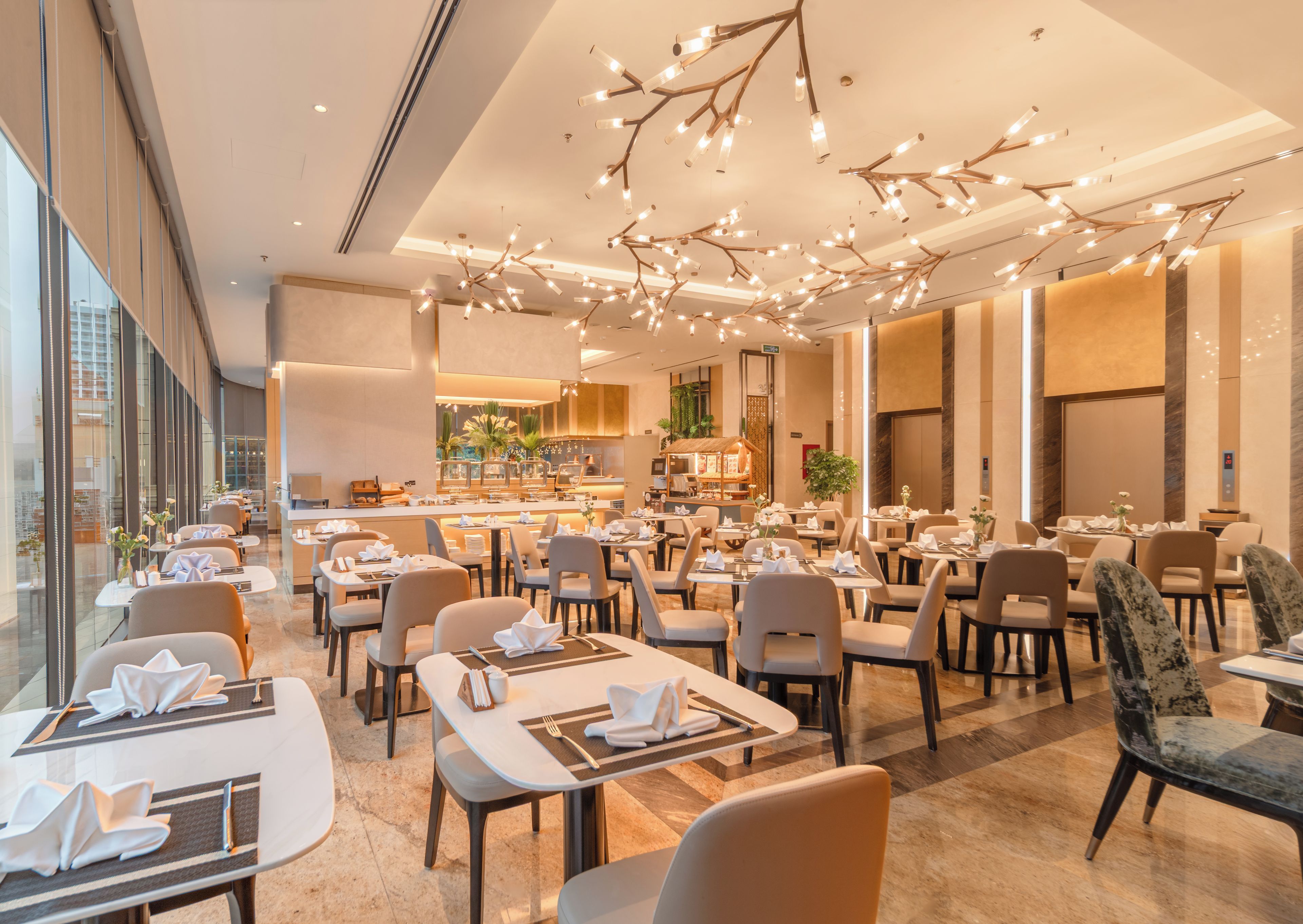 A modern hotel dining area with multiple tables and chairs, a central bar area, and a view of the cityscape through large windows.