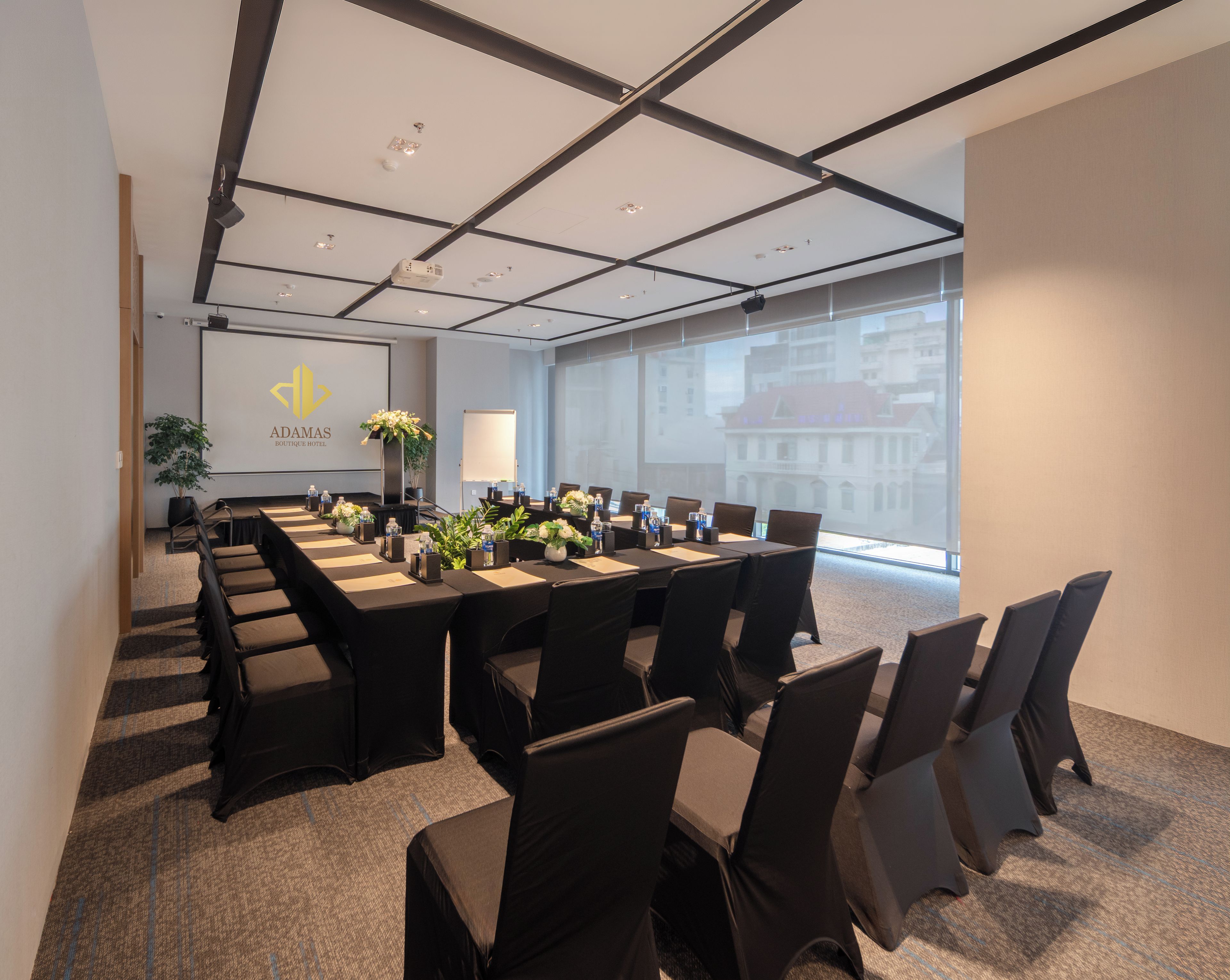 A conference room with a long table, chairs, a projector screen, and a large window with a city view.