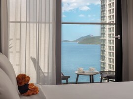 A hotel room with a large window, a bed, a teddy bear, a small table with cups, and a chair.