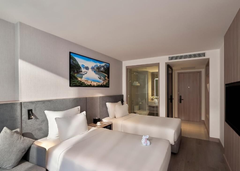 Habitación de hotel con dos camas, un cuadro de un paisaje montañoso, cabina de ducha de vidrio y un televisor.