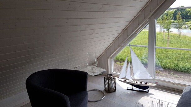 A room with a sloped ceiling, a small sailboat model, a chair, and a window with a view of greenery and a body of water. - Beaver Modern Retreat (Wandlitz)
