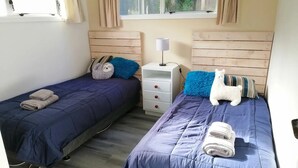 A room with two beds, each with a blue bedspread and pillows, a nightstand with a lamp, and a window with curtains. - 2 bedroom holiday home with outdoor bath & sauna (Upper Hutt)