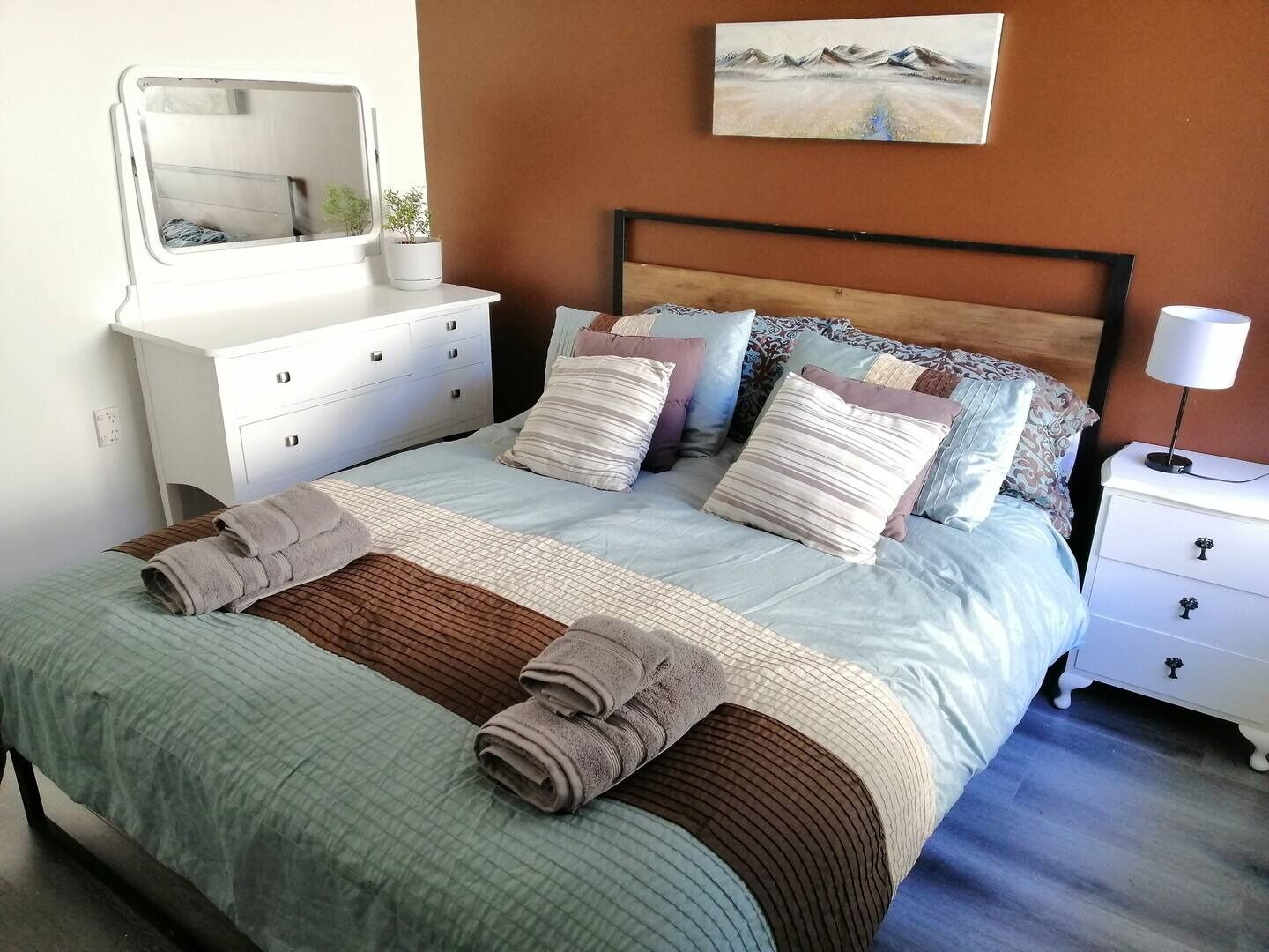 A neatly made bed with a striped blanket and two rolled towels, a bedside table with a lamp, a dresser with drawers, and a painting on the wall.