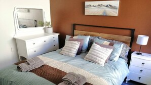 A neatly made bed with a striped blanket and two rolled towels, a bedside table with a lamp, a dresser with drawers, and a painting on the wall. - 2 bedroom holiday home with outdoor bath & sauna (Upper Hutt)