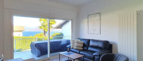 Uma sala de estar moderna com um sofá, uma mesa de centro e uma poltrona. Há uma grande janela com vista para o verde e um edifício.