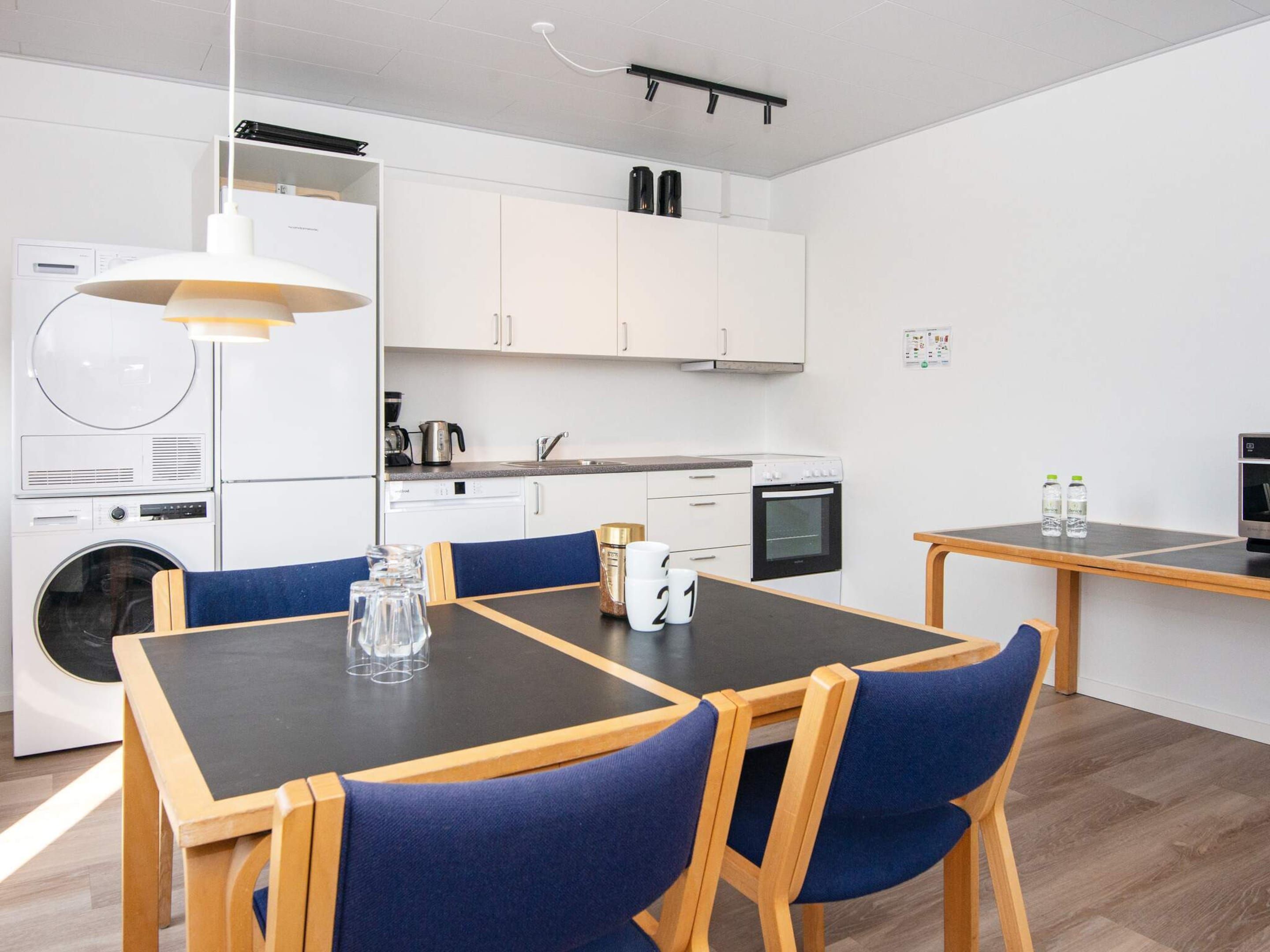 A modern kitchen with white cabinets, a built-in oven, and a coffee machine. There is a dining table with chairs and a hanging light fixture.