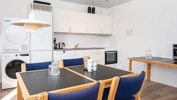 A modern kitchen with white cabinets, a built-in oven, and a coffee machine. There is a dining table with chairs and a hanging light fixture.