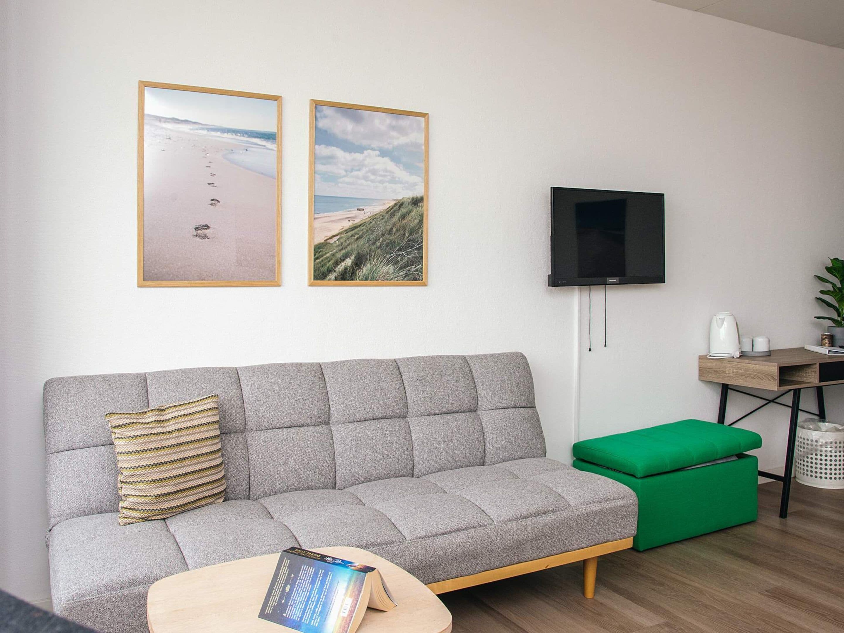 Una sala de estar moderna con un sofá gris, un reposapiés verde, un televisor de pantalla plana y dos cuadros con imágenes de la playa.