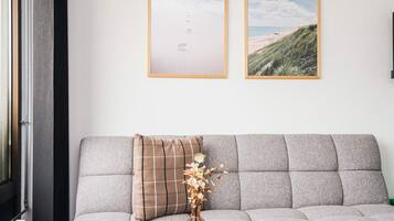 Een grijze bank met een ruitkussen en een vaas met gedroogde bloemen. Boven de bank hangen twee ingelijste foto's.